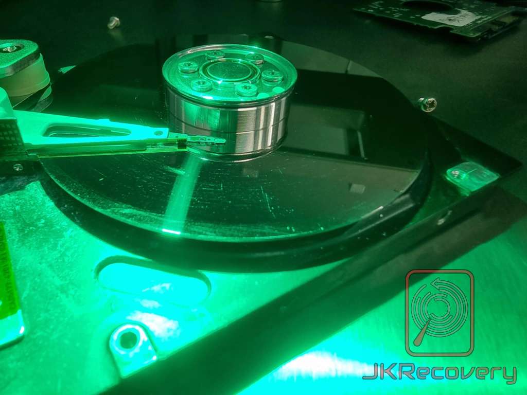 Otwarty dysk twardy HDD oświetlony zielonym światłem UV w laboratorium odzysku danych, widoczne talerze z zarysowaniami powierzchni oraz ramię głowicy z głowicami odczytująco-zapisującymi